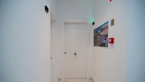 Ein schmaler Flur mit einer weißen Tür am Ende, einem Rauchmelder an der Wand und einem kleinen Plakat.