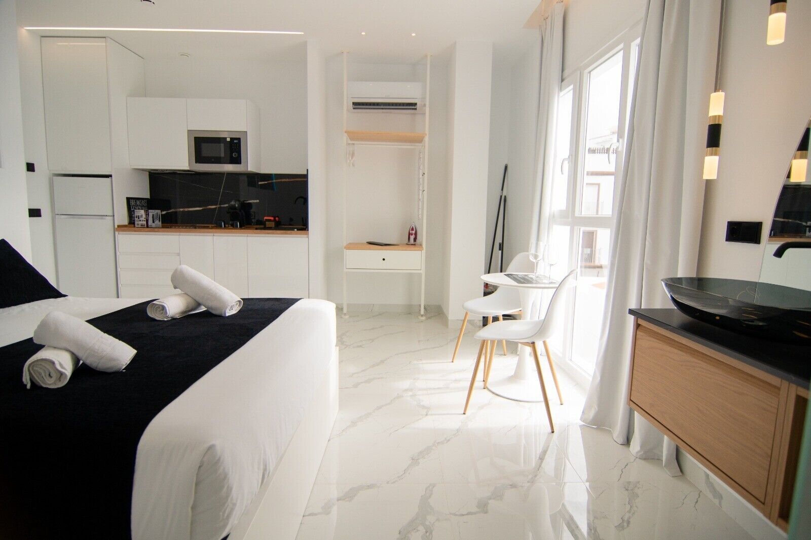 A hotel room with a bed, a desk, a chair, a kitchen area, and a dining table.