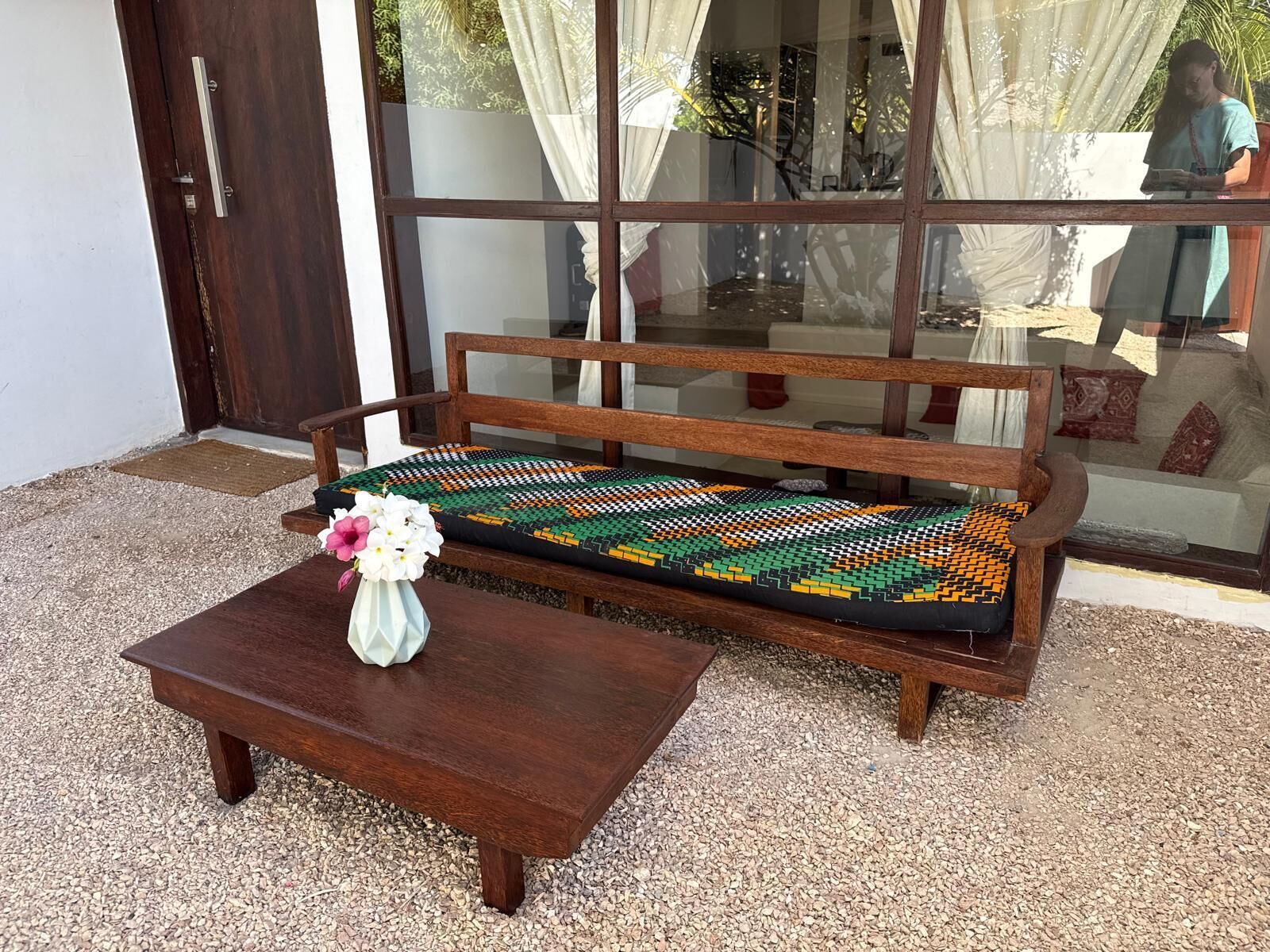 A wooden bench with a patterned cushion, a wooden table with a vase of flowers, and a glass door with curtains.