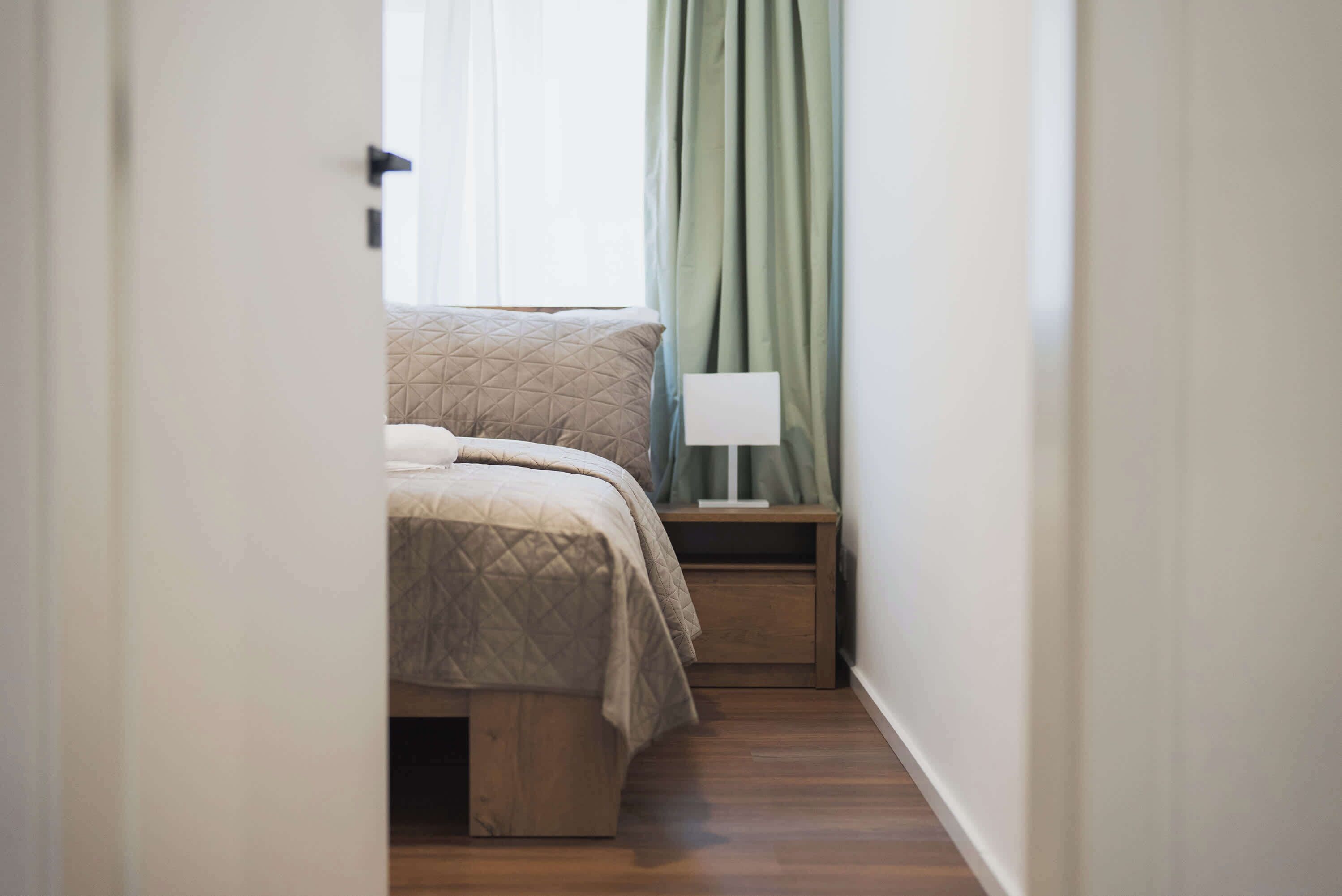 A narrow hallway leading to a bedroom with a bed, a nightstand, and a lamp.