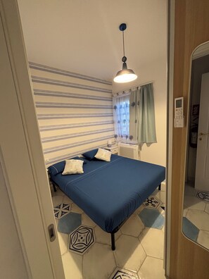 Un dormitorio con una cama, una mesita de noche, una lámpara, una ventana con cortinas y una puerta.