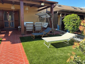 A patio with a brick floor, two wooden chairs, a lounge chair, and a pergola with a hanging light fixture. - Le Tonnare 36 (Sardegna)