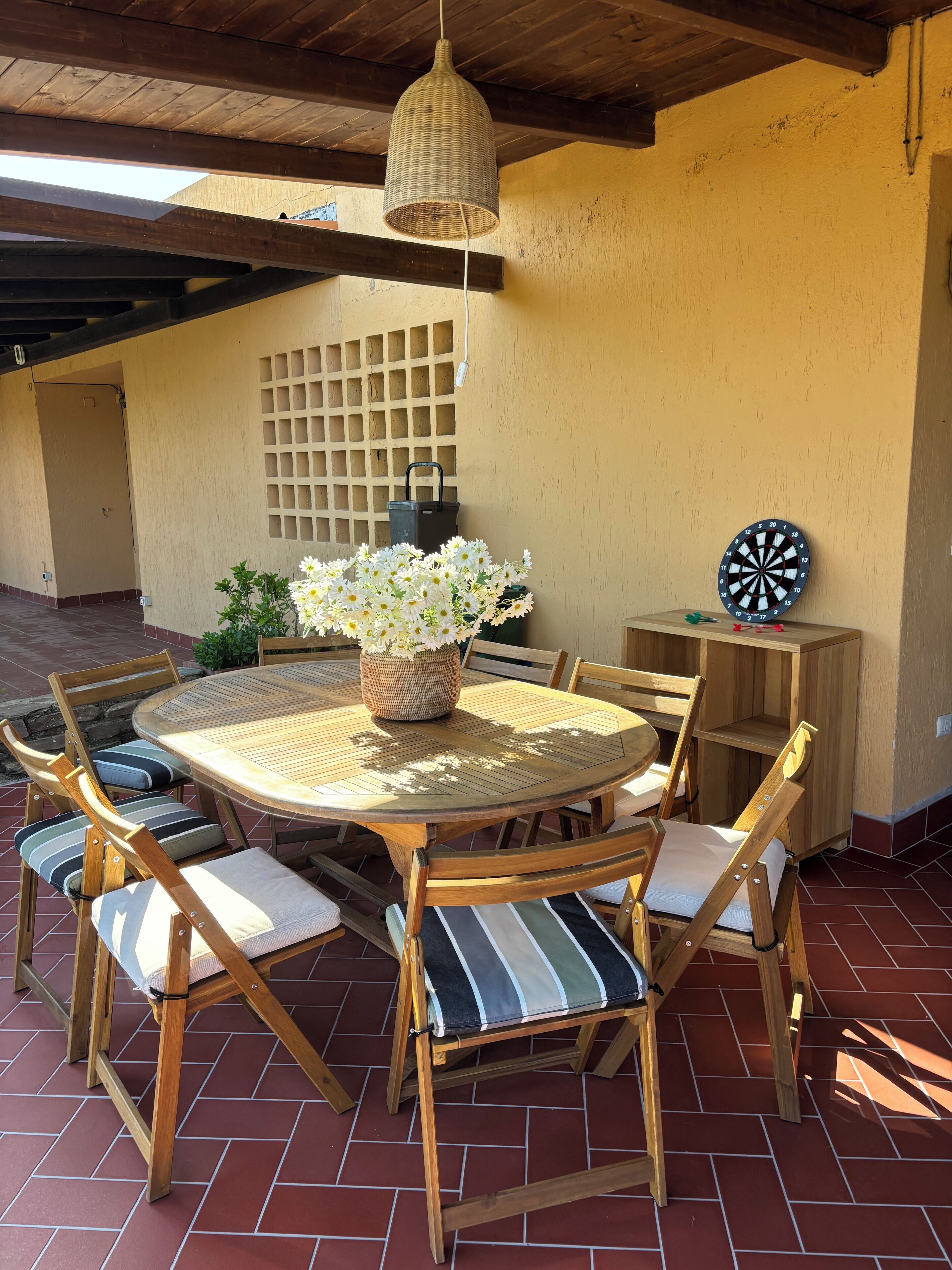 Un patio al aire libre con una mesa redonda de madera, sillas con cojines, una lámpara colgante de fibra natural, una planta en maceta y un dardo.