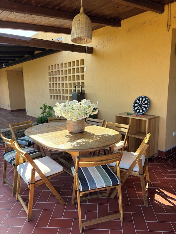 An outdoor patio with a round wooden table, cushioned chairs, a woven hanging light fixture, a potted plant, and a dartboard.