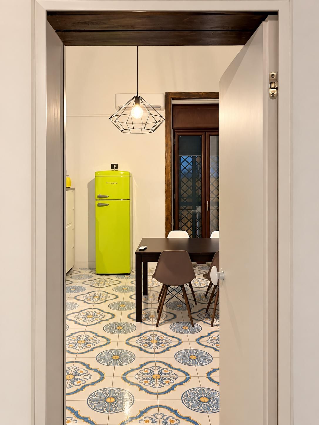 Un couloir étroit mène à une salle à manger où se trouvent une table et des chaises, un réfrigérateur jaune et des carreaux de sol à motifs.