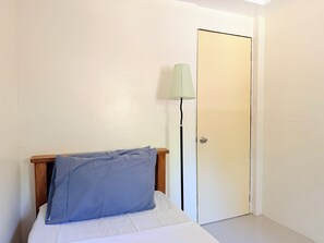 Una cama individual con cabecera de madera y almohada azul, una lámpara de pie y una puerta cerrada.