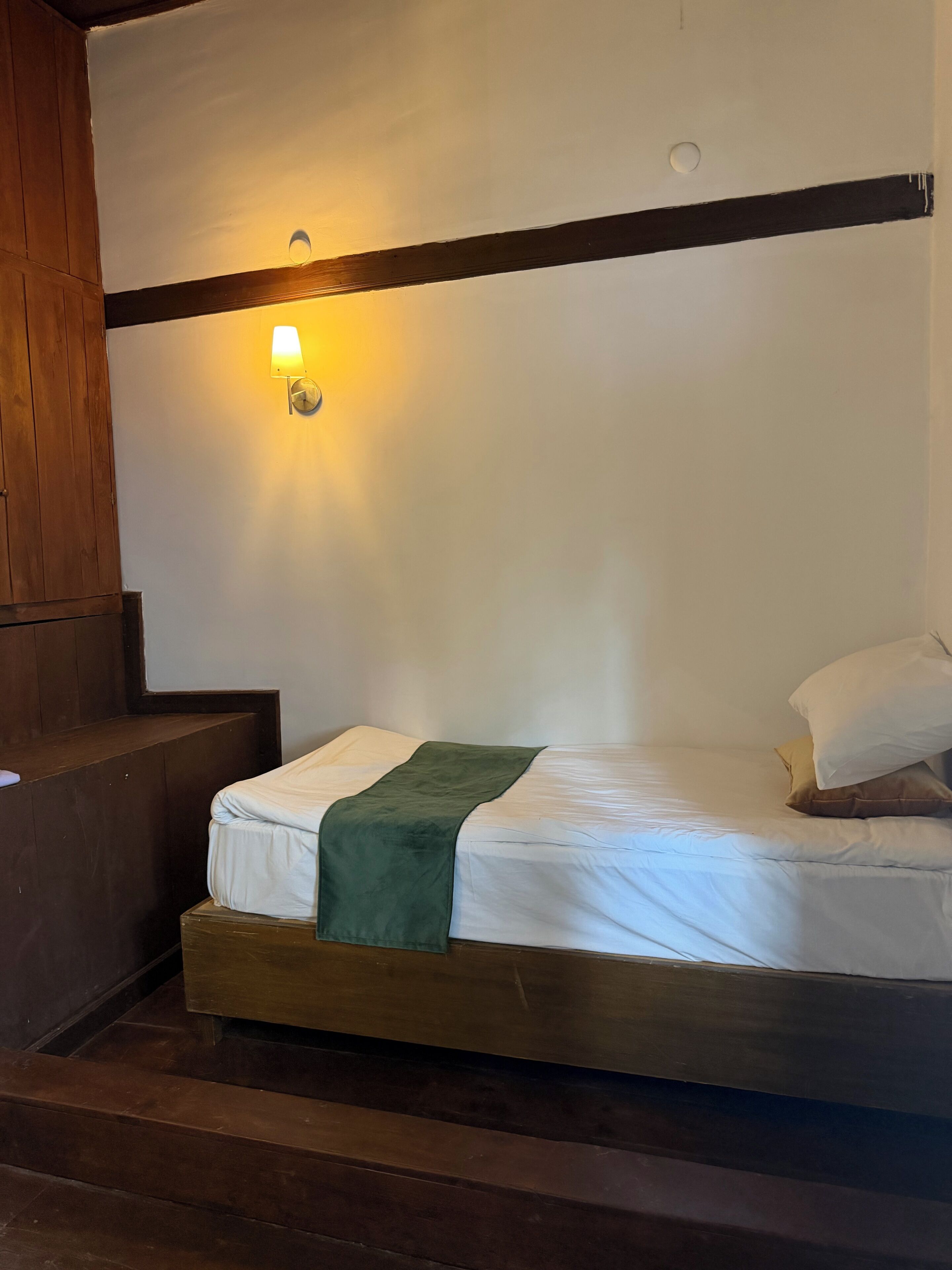 A single bed with a green throw blanket, wooden headboard, and a wall-mounted light fixture.