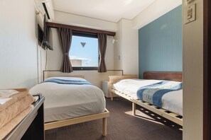 Ein Hotelzimmer mit zwei Einzelbetten, einem an der Wand montierten Fernseher, einem Fenster mit Vorhängen und einem kleinen Tisch mit einem Handtuch.