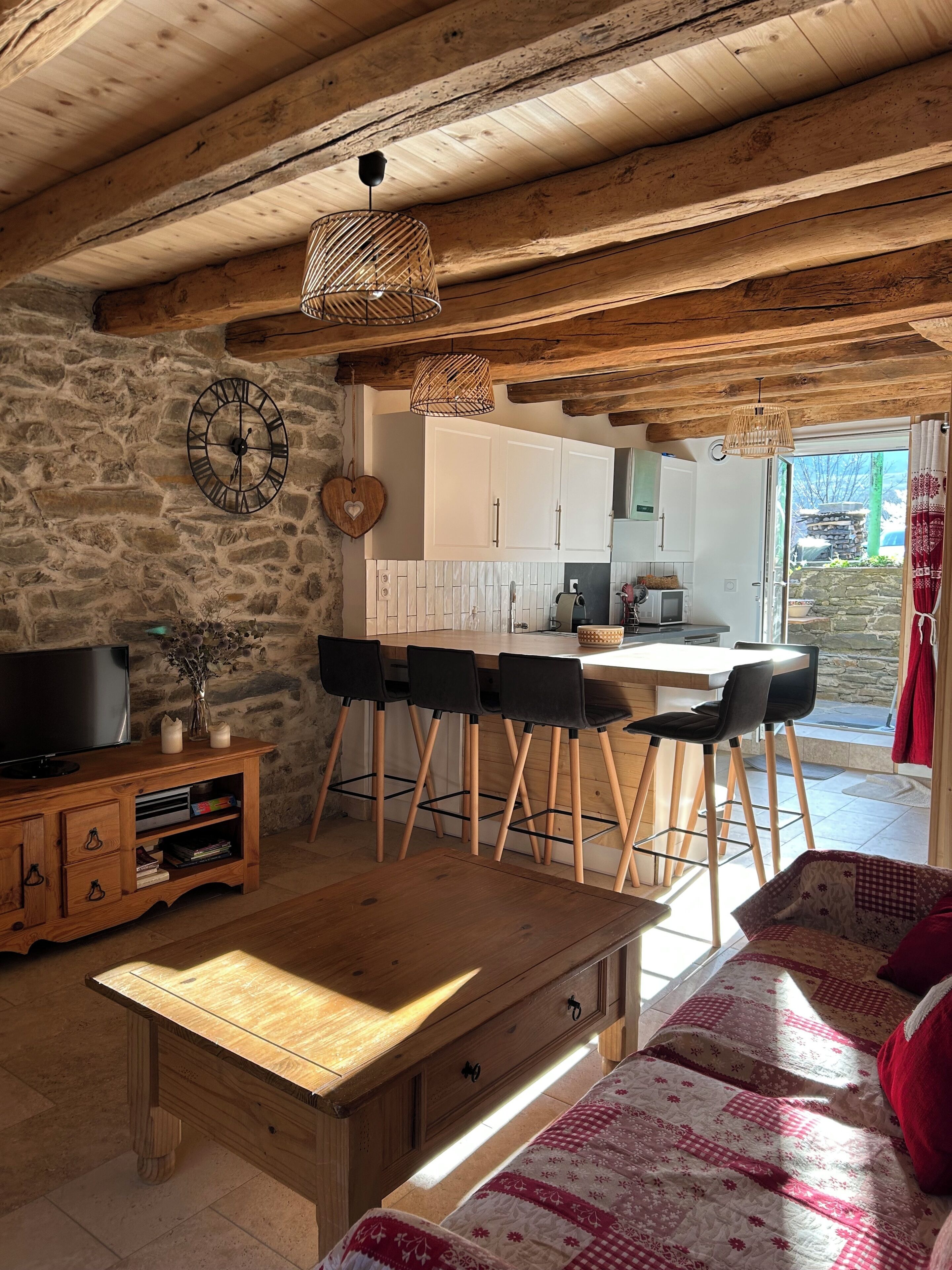 Ein rustikales Interieur mit einer Steinwand, Holzmöbeln, einem Essbereich mit Stühlen, einem Fernseher und einer durchgehenden Tür erkennbaren Küche.