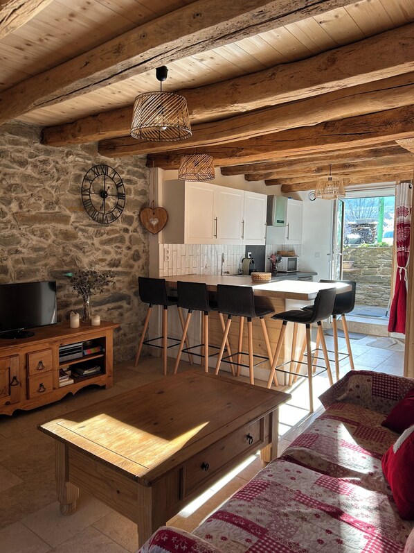 A rustic interior with a stone wall, wooden furniture, a dining area with chairs, a television, and a kitchen visible through an open door. - Apartment with balconies (sleeps 6) (Saint-Michel-de-Maurienne)