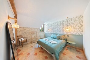 A bedroom with a stone wall, a bed with a blue bedspread, a wooden chair, a hanging light fixture, and a radiator. - A 10-minute drive from Beaune, on a vineyard estate, a charming Burgundian house. (Corcelles-les-Arts)