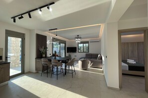 A modern interior with a dining area, kitchen, and living space, featuring a large window, a chandelier, and a flat-screen TV. - Nastazia's Pearl Villa (Afantou)