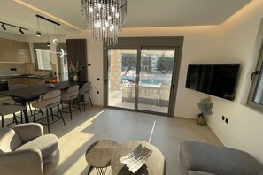 A modern living room with a dining area, a kitchen, and a large sliding glass door leading to an outdoor pool area. - Nastazia's Pearl Villa (Afantou)