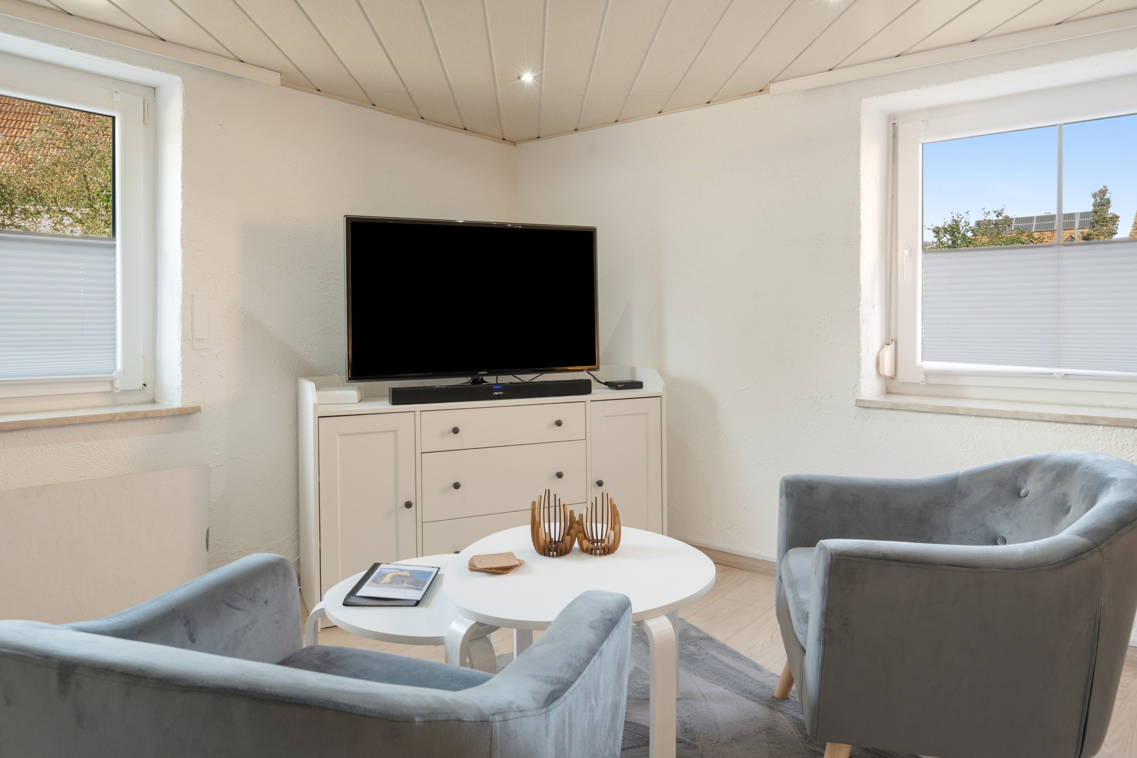 A modern living room with a flat-screen TV, a round white table, two grey armchairs, and a cabinet with drawers.