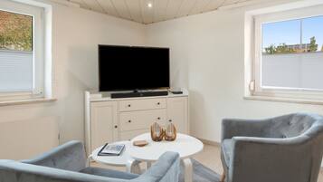 A modern living room with a flat-screen TV, a round white table, two grey armchairs, and a cabinet with drawers.