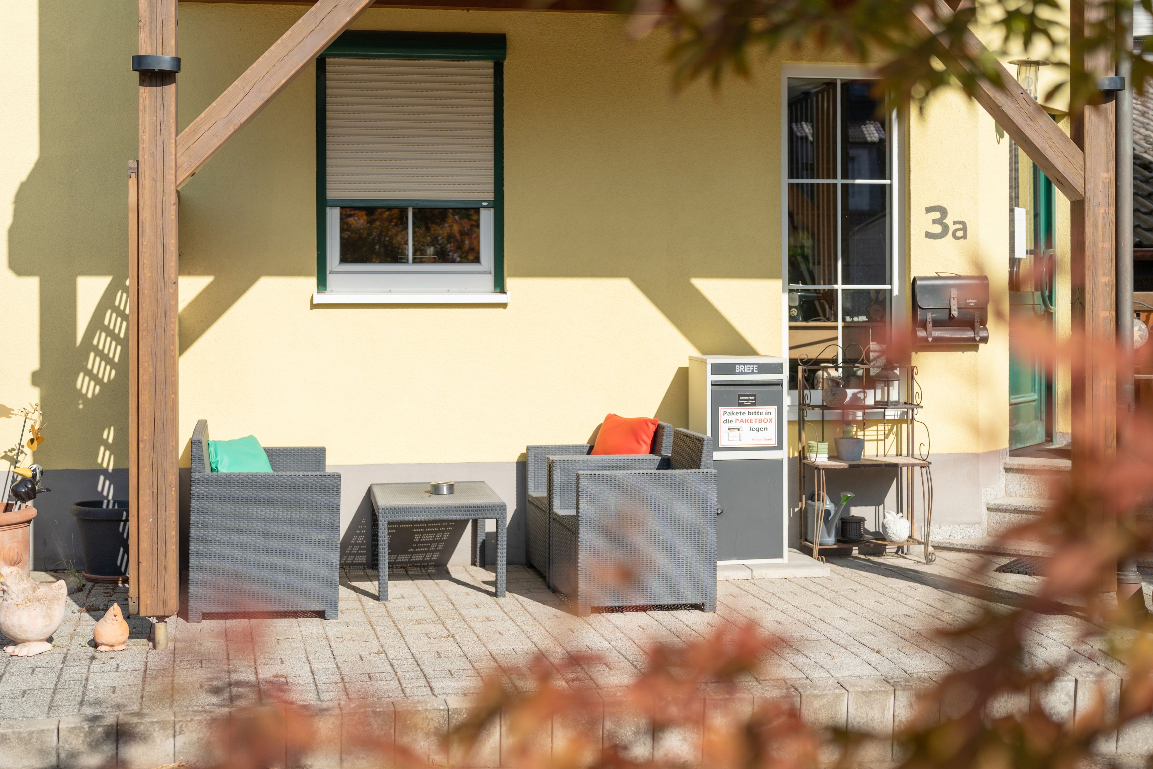 An outdoor seating area with wicker furniture and a small table, a window with blinds, and a door with a mailbox.
