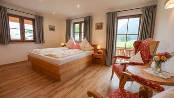 A bedroom with a wooden bed, a nightstand, a chair, a table, and a view of the outdoors through large windows.