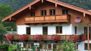 A two-story wooden chalet with a balcony adorned with flowers, surrounded by greenery.