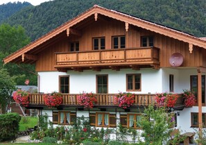 A two-story wooden chalet with a balcony adorned with flowers, surrounded by greenery.