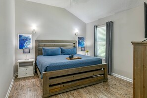 Ein Schlafzimmer mit einem hölzernen Bett, zwei Nachttischen, einem Schreibtischchen, einem Fenster mit Vorhängen und Kunstwerken an den Wänden.