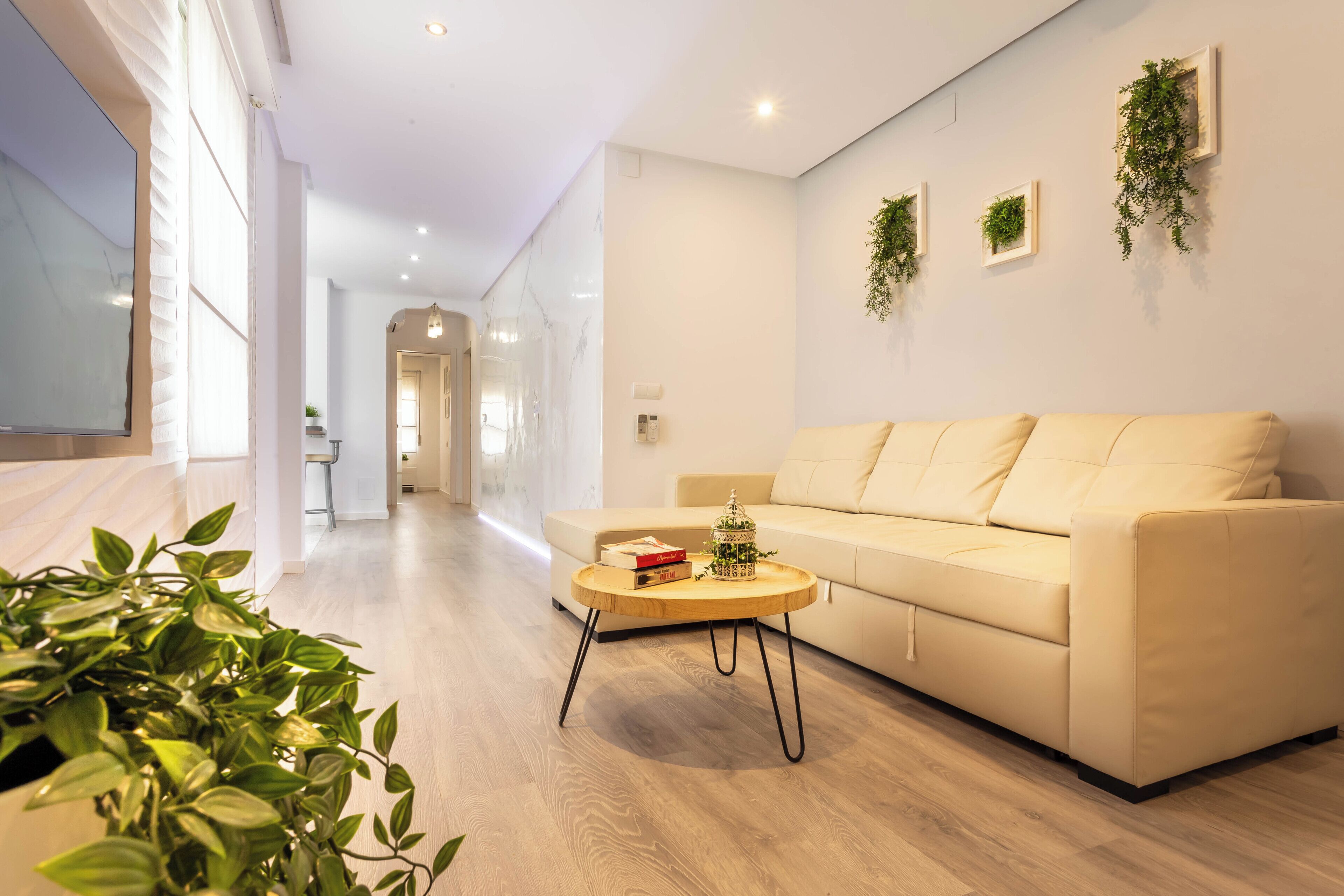 A modern living room with a sofa, a coffee table, and a TV. There are plants on the wall and a hallway leading to another room.