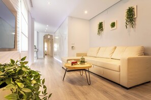 Ein modernes Wohnzimmer mit einer Couch, einem Couchtisch und einem Fernseher. An der Wand befinden sich Pflanzen und ein Flur, der in einen anderen Raum führt.