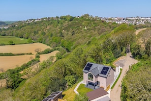 A modern house with solar panels, surrounded by lush greenery and fields.
