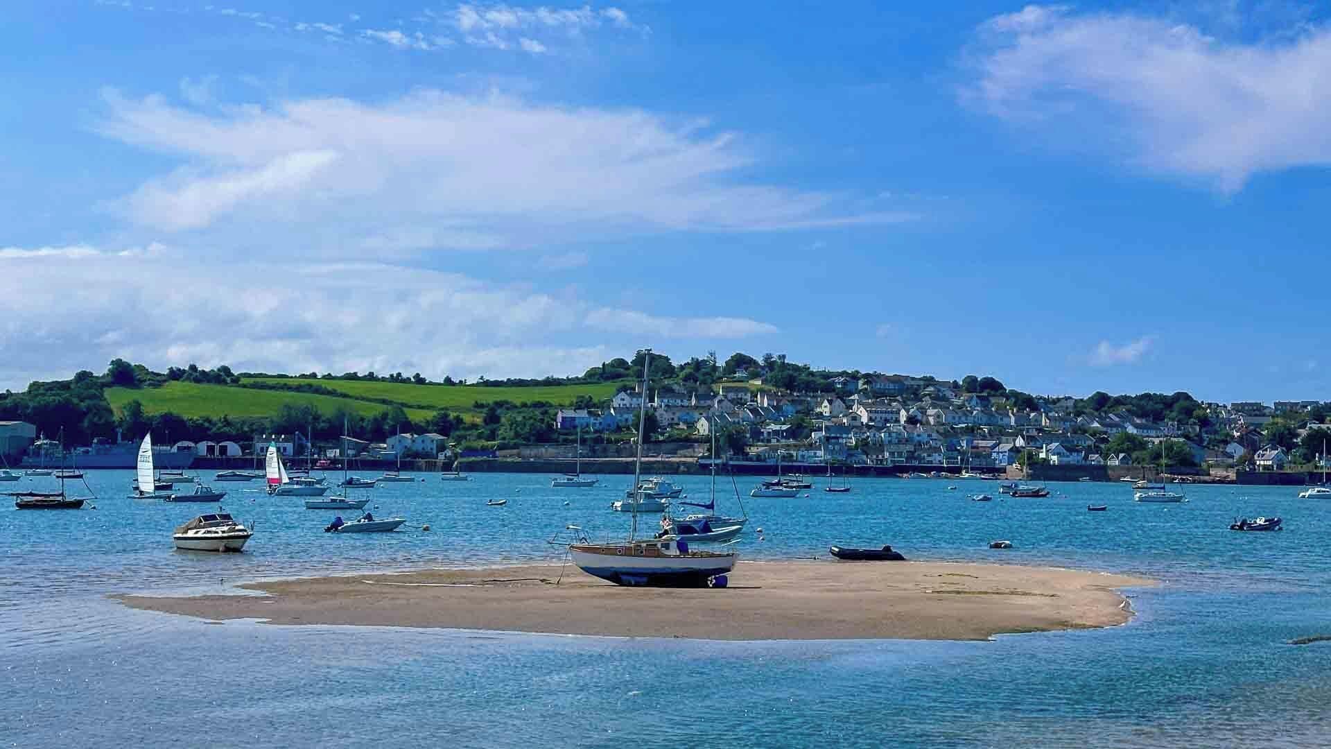 A coastal scene with boats on calm waters, a sandy beach, and a town in the background.