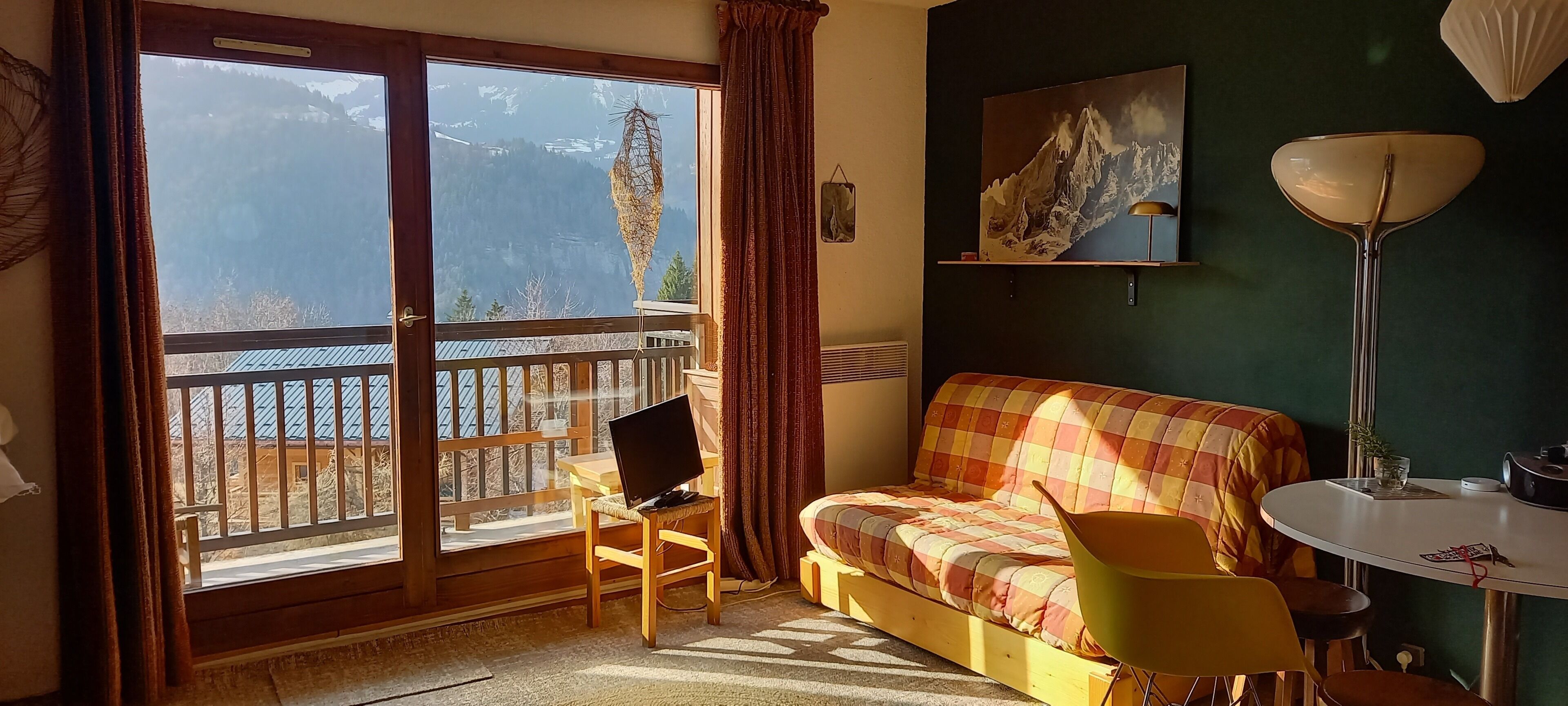 Een kamer met balkon, een bed met geruit dekbed, een stoel, een tafel en een lamp.