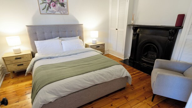 A bedroom with a bed, bedside tables, a chair, a fireplace, and a painting on the wall.