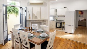 A dining area with a wooden table and white chairs, a kitchen with white cabinets and appliances, and a view of the outdoors through a glass door.