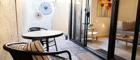 Eine Terrasss mit runden Tisch und zwei Stühlen, eine Glastür zu einem Innenraum mit einer Couch und einem Gemälde.