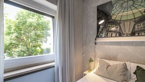 Un dormitorio con cama, mesita de noche, lámpara, ventana con cortinas y un mural decorativo en la pared.