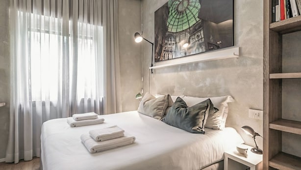 Habitación de hotel con cama, mesita de noche, lámpara, un cuadro y ventana con cortinas.