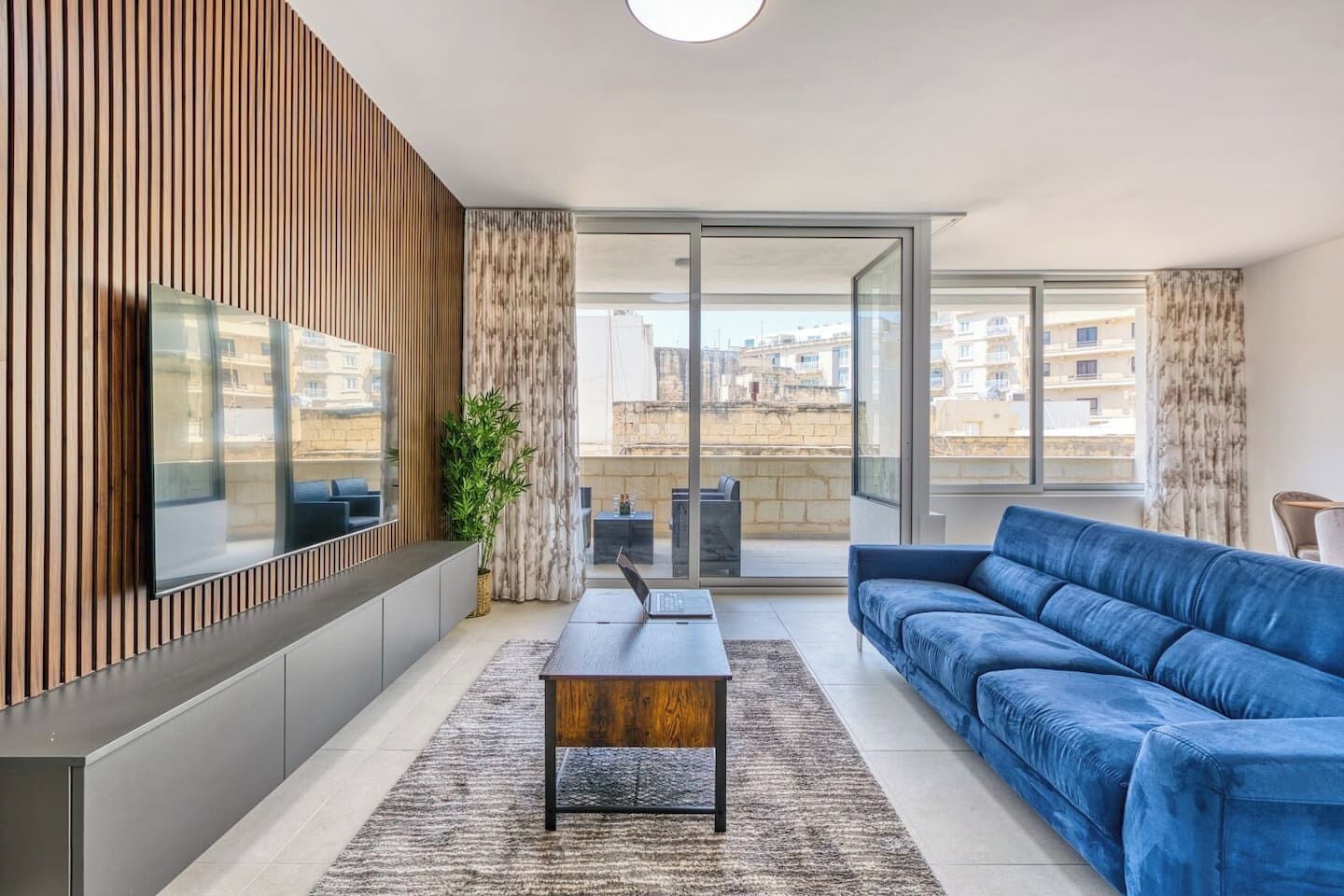 En moderne stue med sofa, sofabord, tv og en balkon med udsigt til bygninger.