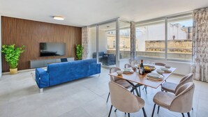 A modern living room with a blue sofa, a dining table set for four, and a TV mounted on a wooden panel wall. - Beautiful, fully equipped 3BR in Sliema w/Balcony (Tas-Sliema)