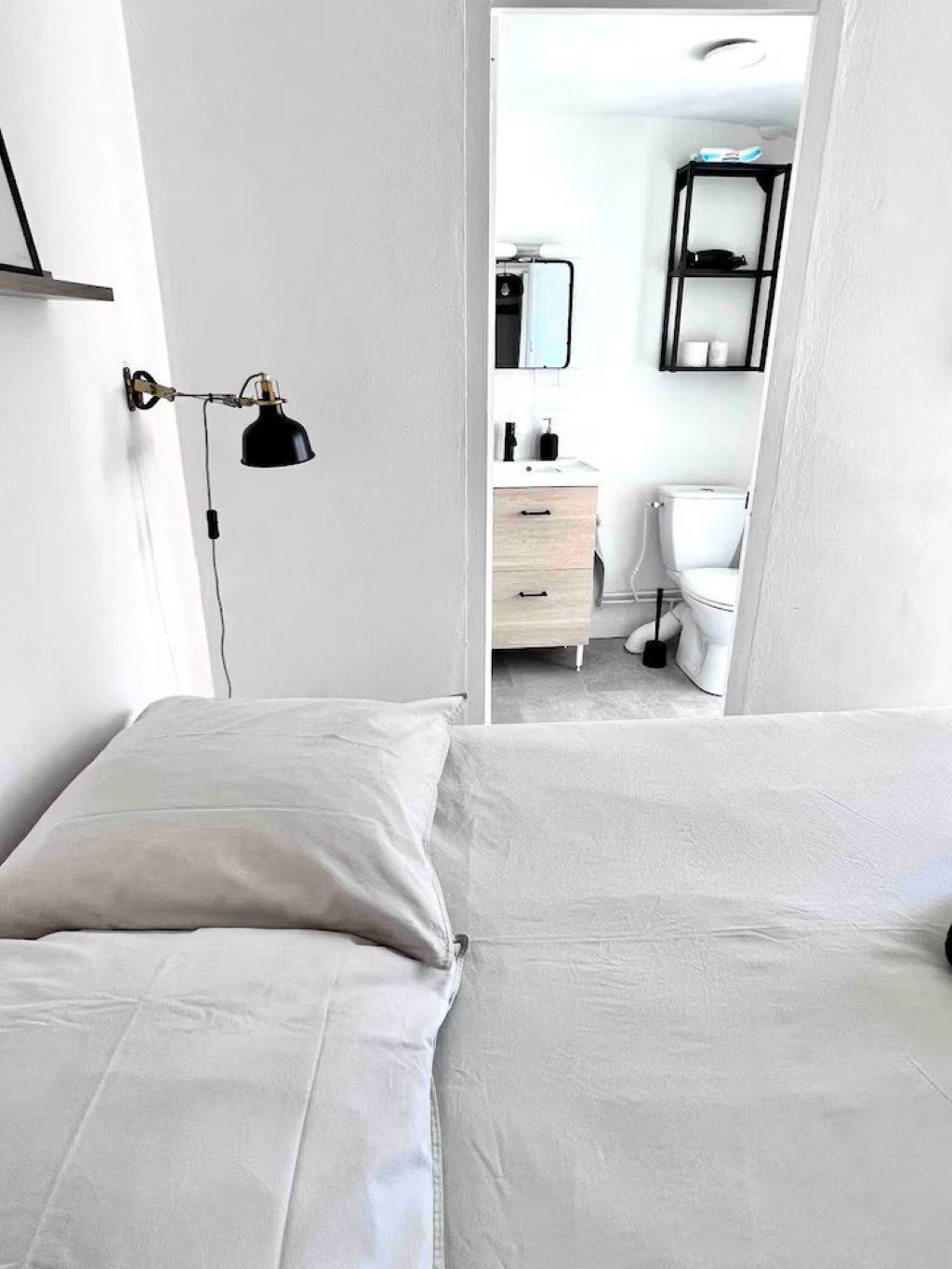 A bedroom with a bed, a nightstand, a wall-mounted lamp, and a view into a bathroom with a toilet and a shelf.