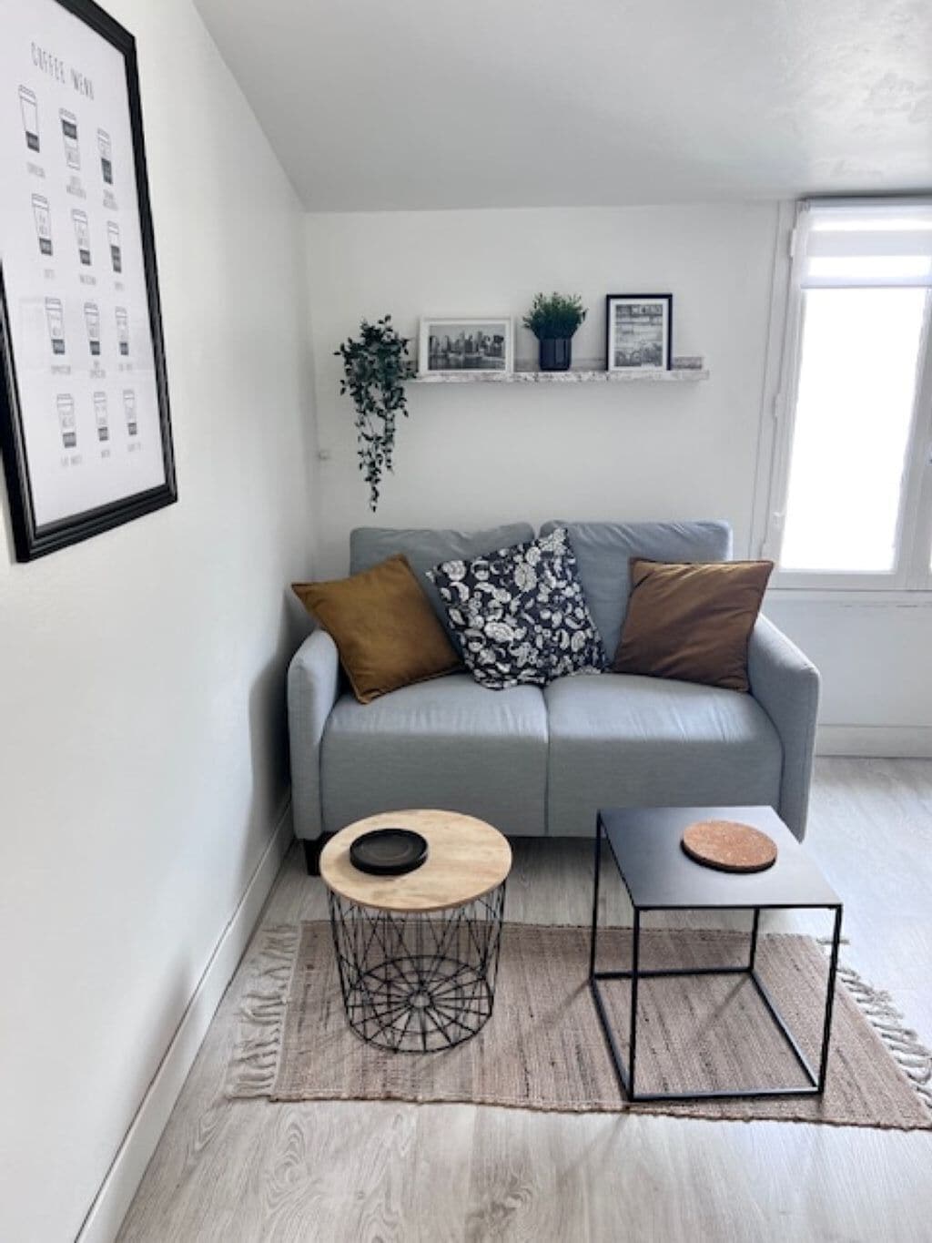 A modern living room with a grey sofa, two side tables, a coffee menu on the wall, and a window with blinds.
