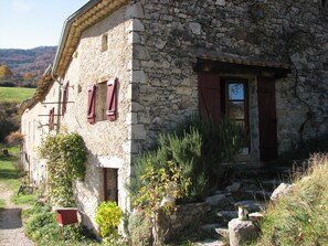 Ein Steinhaus mit Tür und Fenstern, umgeben von Pflanzen und einem Pfad.