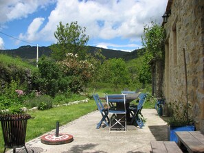 Un espacio al aire libre con mesa y sillas, rodeado de follaje y un muro de piedra.