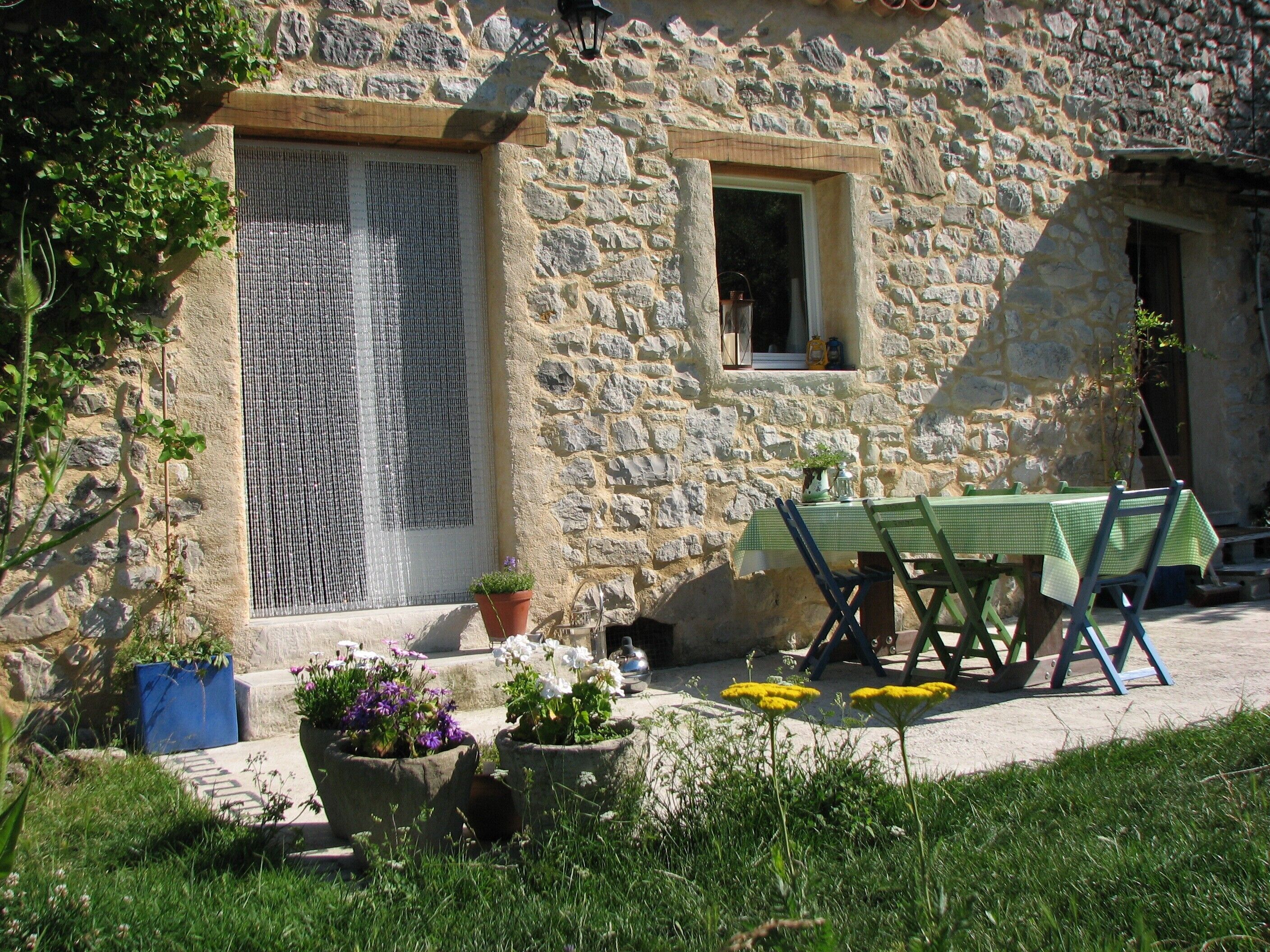 A stone building with a window, a door, and a table with chairs outside. There are potted plants and flowers in front.