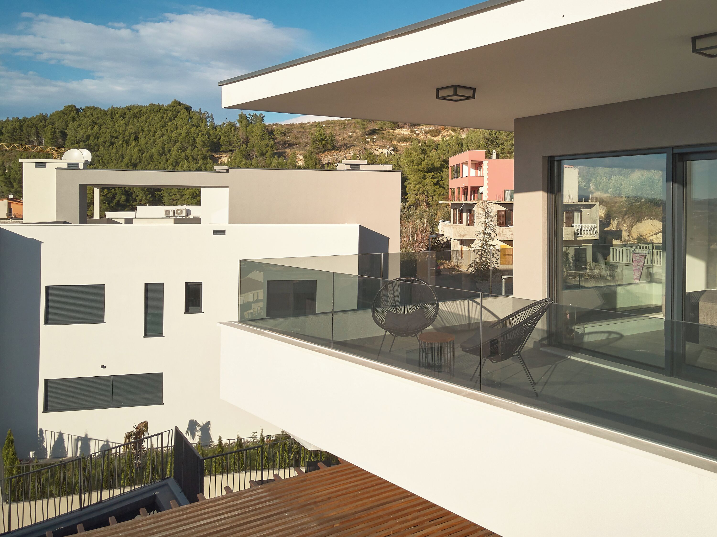 A modern balcony with a glass railing, a wooden deck, and a view of a hillside with buildings.