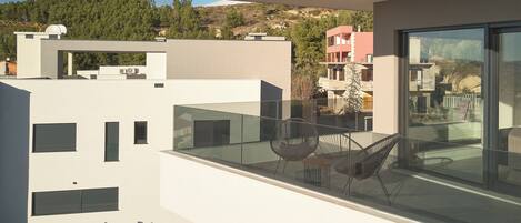 A modern balcony with a glass railing, a wooden deck, and a view of a hillside with buildings.