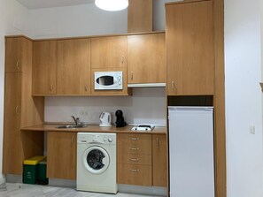 Eine kleine Küche mit Holzschränken, einer Mikrowelle, einer Waschmaschine, einem Wasserkocher und einem Kühlschrank.