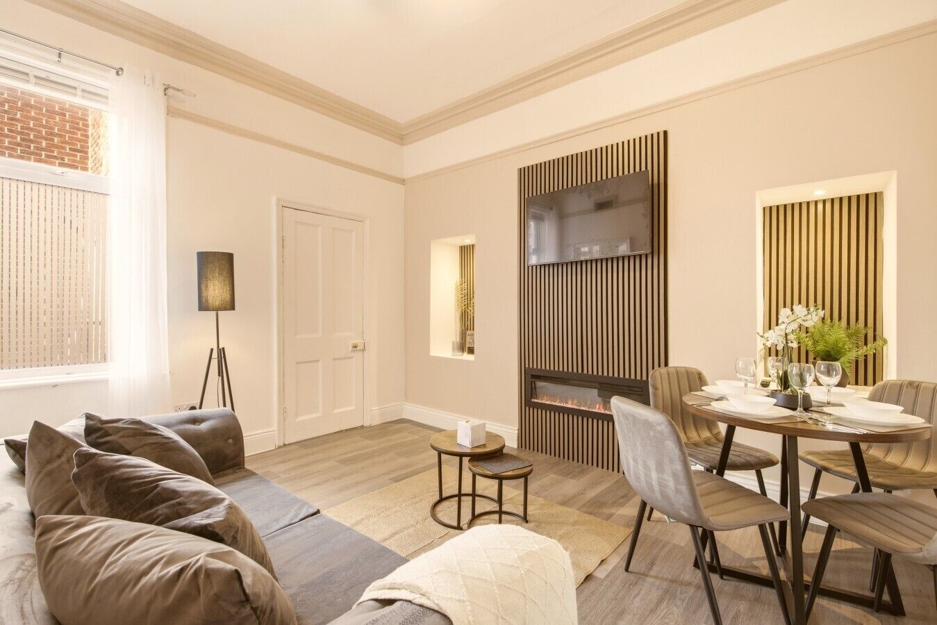 A modern living room with a sofa, a dining table set for four, a fireplace, and a television mounted on the wall.
