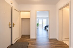 A hallway with a wooden floor, white walls, and a door leading to a room with a plant and a sofa.