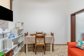Un petit coin repas avec une table et des chaises, un téléviseur placé au-dessus de la table et un canapé agrémenté d’un coussin coloré.