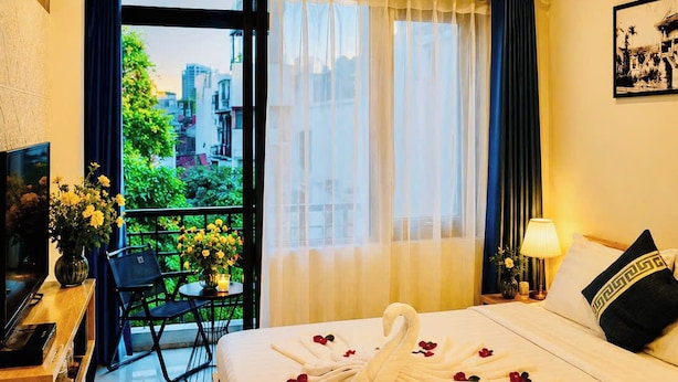 A hotel room with a bed, a TV, a desk with a vase of flowers, a balcony with a view of buildings, and a window with sheer curtains. - Silver Lantern Hanoi Hotel (Hanoi)