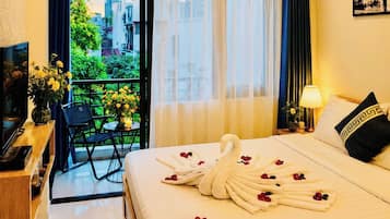 A hotel room with a bed, a TV, a desk with a vase of flowers, a balcony with a view of buildings, and a window with sheer curtains.