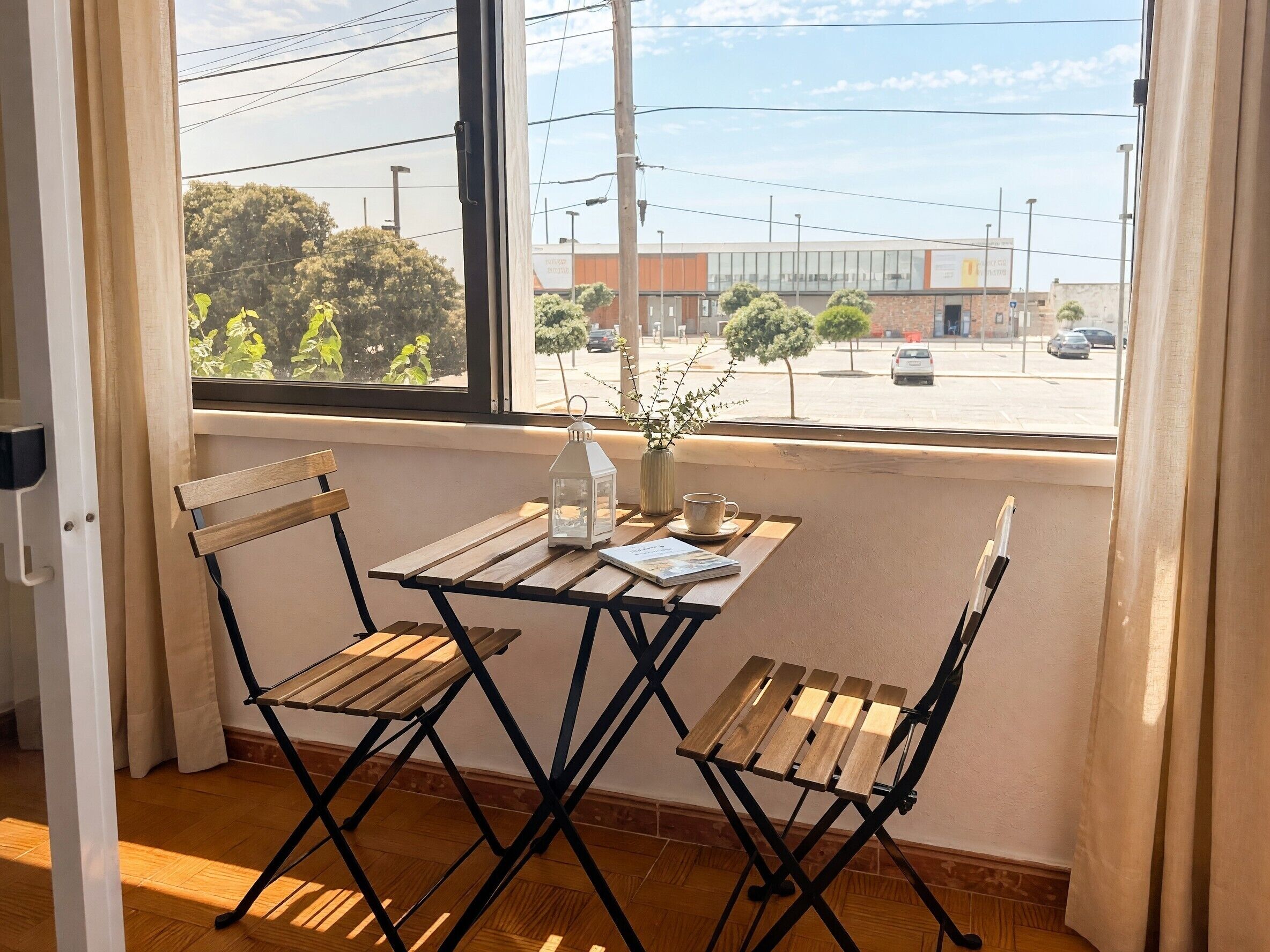 Una habitación con vista a un estacionamiento y edificios, una pequeña mesa con un libro y una vela, y dos sillas de madera.